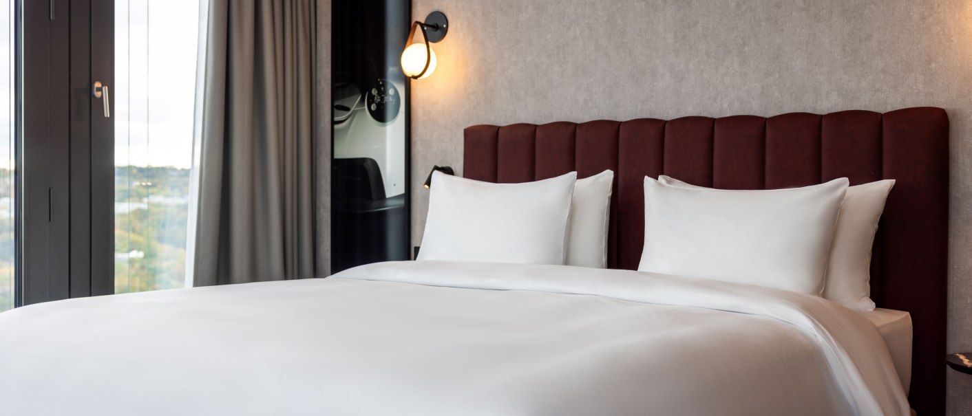 Ein elegantes Hotelzimmer mit einem großen Bett, weißen Bettlaken und einem roten Kopfteil. Links befindet sich ein Fenster mit grauen Vorhängen., © Radisson Hotel Group