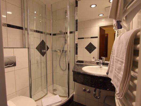 Modernes Badezimmer mit Glasdusche, Waschbecken und Handtuchhalter. Weiße Fliesen mit schwarzen Akzenten. Beleuchtung an der Decke., © Aparthotel Astro