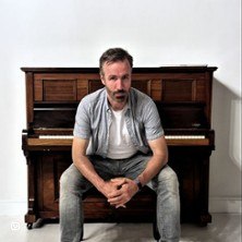 Ein Mann sitzt vor einem Klavier und blickt ernst in die Kamera. Er tr&auml;gt ein wei&szlig;es T-Shirt und eine offene Hemdjacke., &copy; links im Bild