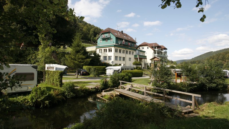 Ein Resort mit Wohnwagen, Gebäuden und einer Holzbrücke in einer grünen, bewaldeten Landschaft unter blauem Himmel., © Family Resort Kleinenzhof Blick in den Nord Schwarzwald
