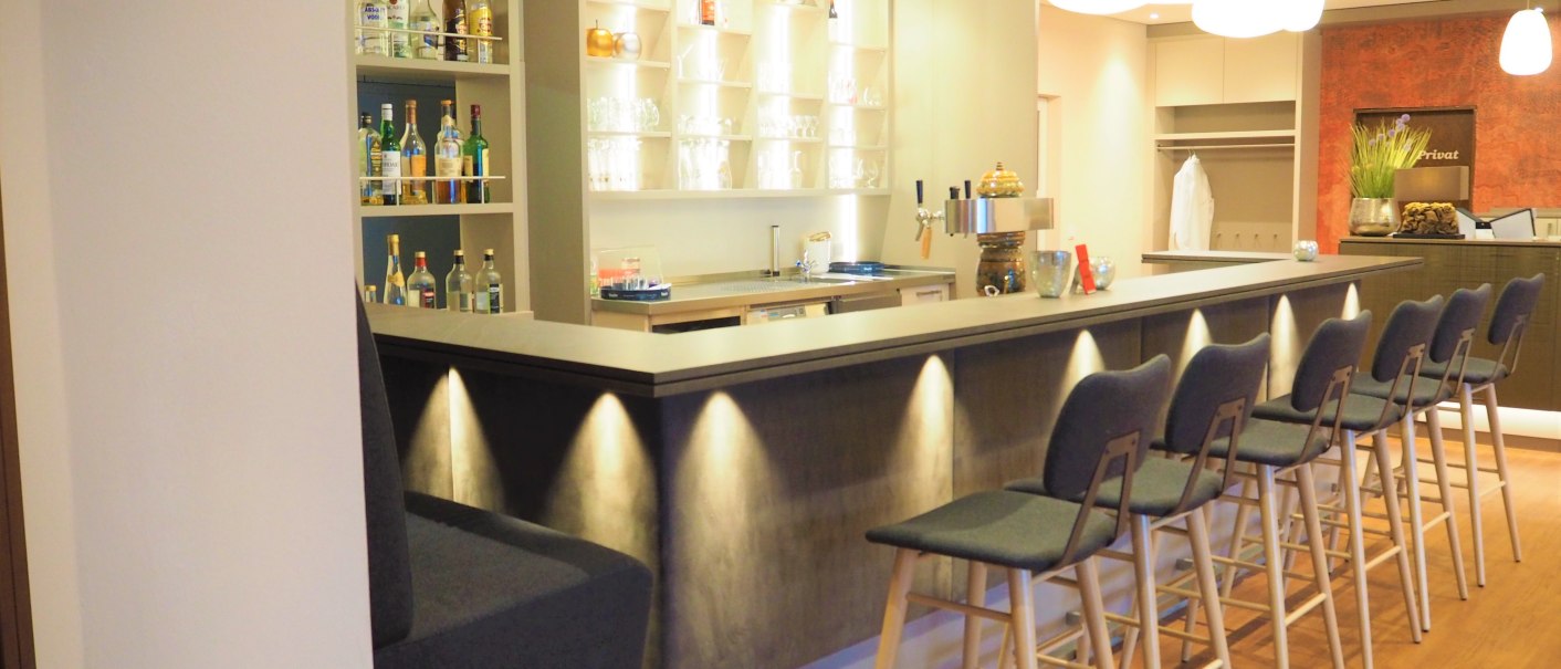 Eine moderne Bar mit hohen Stühlen, beleuchtetem Tresen und Regalen voller Flaschen. Die Beleuchtung ist warm und einladend., © TOMAS Eine moderne Bar mit hohen Stühlen, beleuchtetem Tresen und Regalen voller Flaschen. Die Beleuchtung ist warm und einladend., © TOMAS