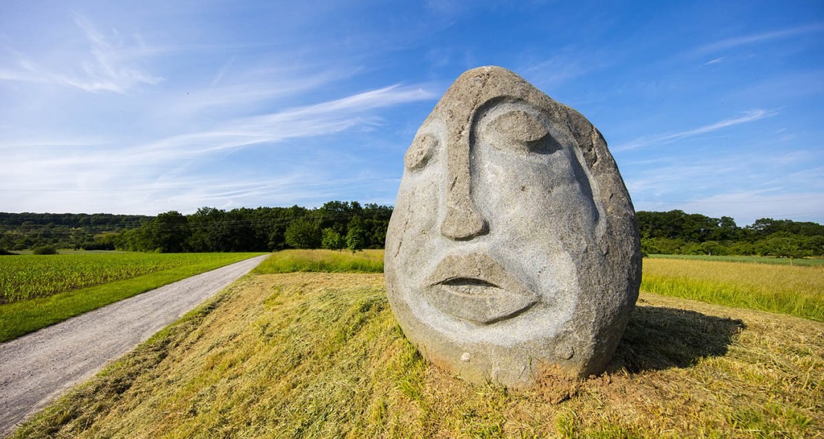 Große Steinskulptur eines Gesichts auf einem Feldweg, umgeben von grünen Feldern und blauem Himmel., © Remstal Tourismus e.V. Große Steinskulptur eines Gesichts auf einem Feldweg, umgeben von grünen Feldern und blauem Himmel., © Remstal Tourismus e.V.