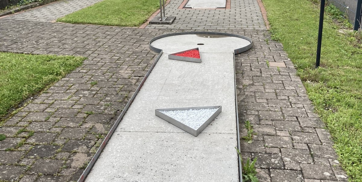 Minigolfbahn mit bunten Dreiecken als Hindernisse auf einem gepflasterten Weg, umgeben von Bäumen und Rasen.