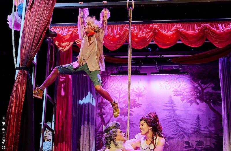 Theaterszene aus 'Der eingebildete Kranke' mit drei Schauspielern, einer hängt an einem Seil, die anderen sitzen darunter. Bunte Kulisse mit roten Vorhängen., © Stadt Kornwestheim Kulturmanagement