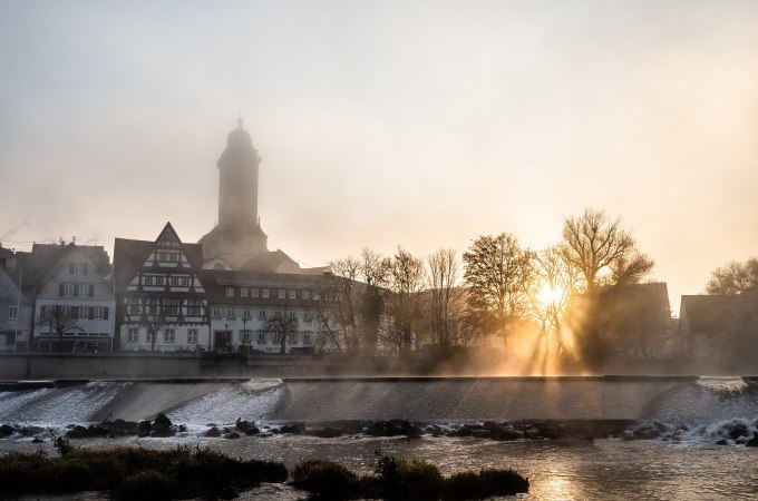 Nebelverhangene Stadtansicht von N&uuml;rtingen mit Fachwerkh&auml;usern und Kirchturm im Morgenlicht. Die Sonne bricht durch die B&auml;ume am Fluss., &copy; Stadt N&uuml;rtingen