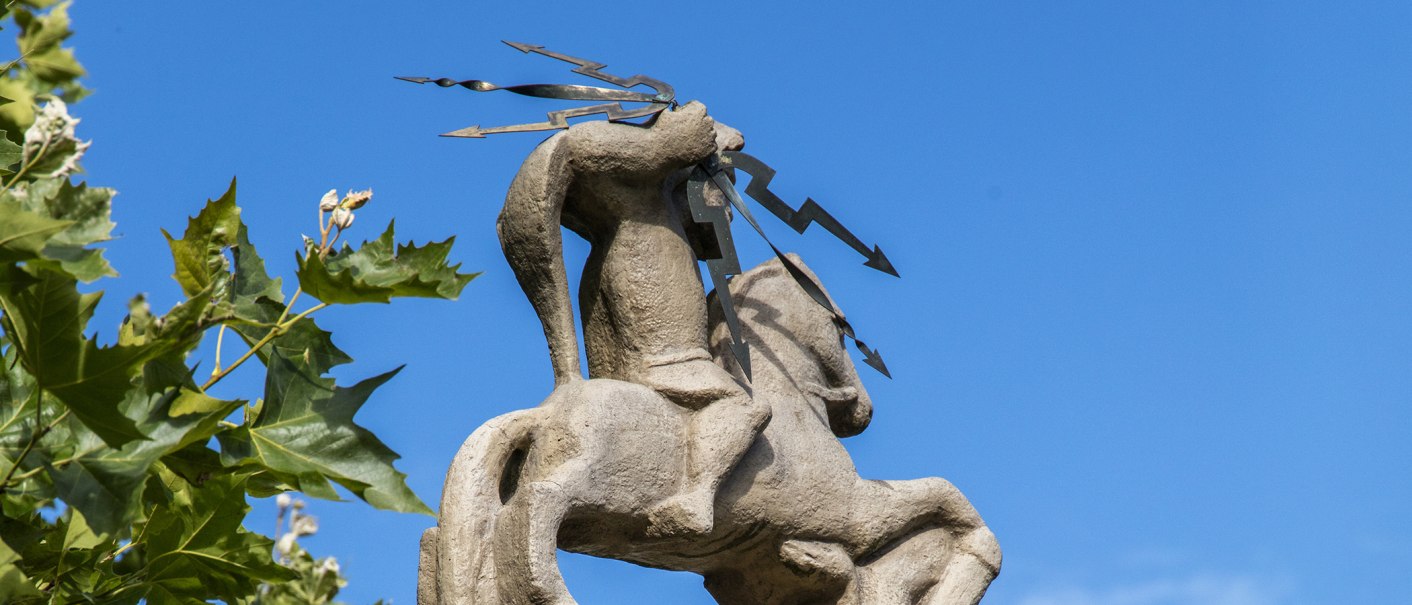 Steinfigur eines Reiters mit Blitzsymbolen, umgeben von grünen Blättern, vor klarem, blauem Himmel., © SMG, Achim Mende Steinfigur eines Reiters mit Blitzsymbolen, umgeben von grünen Blättern, vor klarem, blauem Himmel., © SMG, Achim Mende