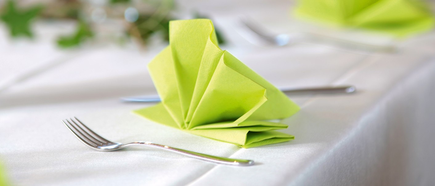 Elegante Tischdekoration mit gefalteter gr&uuml;ner Serviette und Besteck auf wei&szlig;er Tischdecke. Im Hintergrund sind unscharfe gr&uuml;ne Bl&auml;tter zu sehen., &copy; Landgasthof Linde