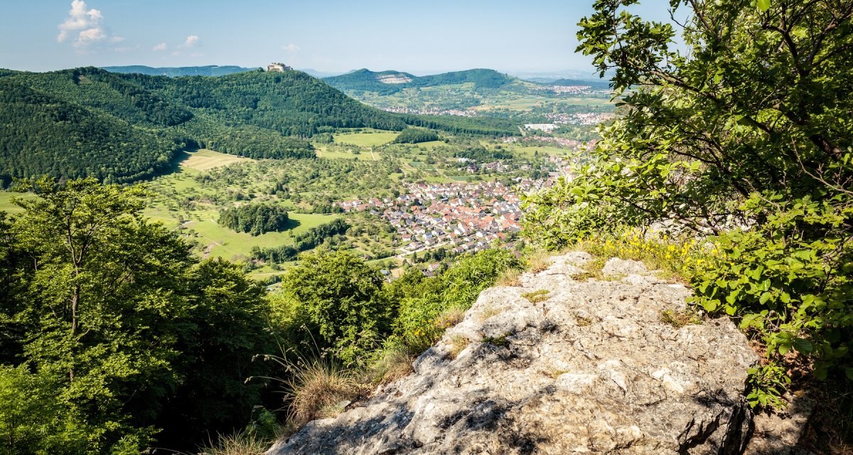 Blick auf eine grüne Landschaft mit Hügeln, einem Dorf und der Burg Hohenneuffen in der Ferne. Im Vordergrund Felsen und Bäume., © hochgehberge Blick auf eine grüne Landschaft mit Hügeln, einem Dorf und der Burg Hohenneuffen in der Ferne. Im Vordergrund Felsen und Bäume., © hochgehberge