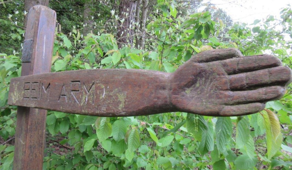 Holzskulptur eines ausgestreckten Arms mit der Inschrift 'BEIM ARM' vor grünem Laub im Wald., © Natur.Nah. Schönbuch & Heckengäu Holzskulptur eines ausgestreckten Arms mit der Inschrift 'BEIM ARM' vor grünem Laub im Wald., © Natur.Nah. Schönbuch & Heckengäu