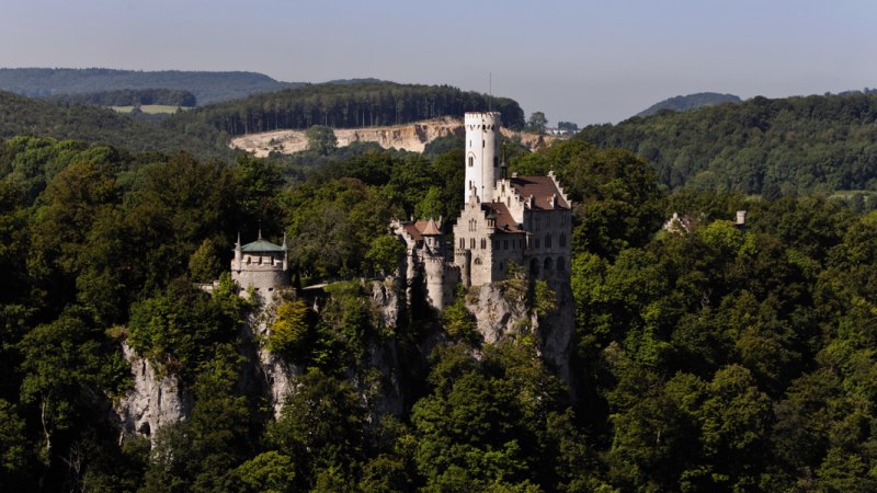 Schloss Lichtenstein thront majestätisch auf einem bewaldeten Hügel, umgeben von dichtem Wald und sanften Hügeln im Hintergrund., © Stuttgart-Marketing GmbH