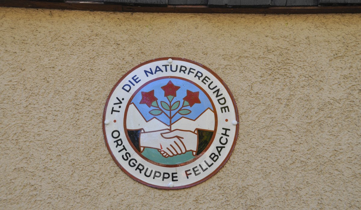Rundes Logo der Naturfreunde Ortsgruppe Fellbach auf einer beigen Wand. Es zeigt zwei Hände, die sich schütteln, und drei rote Blumen vor Bergkulisse. Rundes Logo der Naturfreunde Ortsgruppe Fellbach auf einer beigen Wand. Es zeigt zwei Hände, die sich schütteln, und drei rote Blumen vor Bergkulisse.
