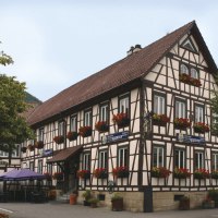Fachwerkhaus mit roten Blumen an den Fenstern, Restaurant im Erdgeschoss, umgeben von Bäumen und einem sonnigen Himmel.