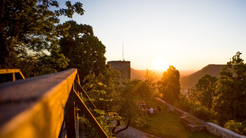 Die Burgruine Helfenstein bei Sonnenuntergang, umgeben von Bäumen. Menschen sitzen auf einer Wiese und genießen die Aussicht., © Arrondissement de Göppingen Die Burgruine Helfenstein bei Sonnenuntergang, umgeben von Bäumen. Menschen sitzen auf einer Wiese und genießen die Aussicht., © Arrondissement de Göppingen
