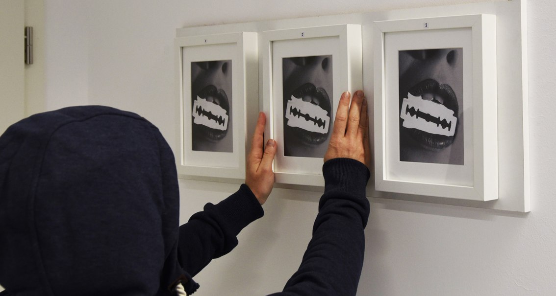 Person in Kapuzenpullover hängt drei gerahmte Bilder mit Lippen und Rasierklingen an eine weiße Wand., © Flucht-Raum