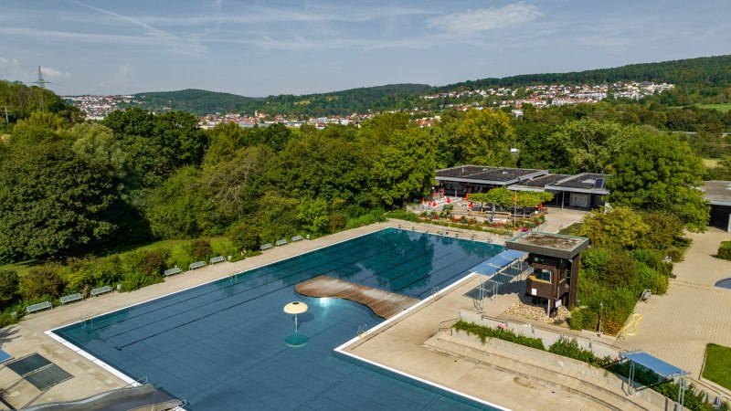 Luftaufnahme des Reichenbacher Freibads, umgeben von grünen Bäumen und Hügeln. Ein großes Schwimmbecken und Gebäude sind sichtbar., © Gemeinde Reichenbach a.d. Fils, Ingo Rack