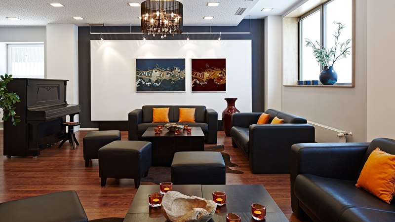 Elegante Lounge mit schwarzen Sofas, orangefarbenen Kissen, Klavier, Kunstwerken und Kerzen auf Holztischen., © Studierendenwerk Stuttgart Elegante Lounge mit schwarzen Sofas, orangefarbenen Kissen, Klavier, Kunstwerken und Kerzen auf Holztischen., © Studierendenwerk Stuttgart