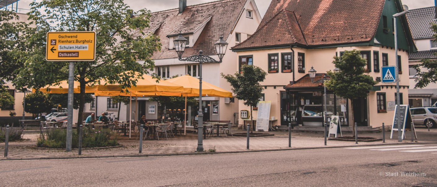 Gemütliche Straßenszene mit Fachwerkhäusern, gelben Sonnenschirmen und einem Café. Ein Wegweiser zeigt Richtungen an. Blätter hängen im Vordergrund., © Stadt Welzheim