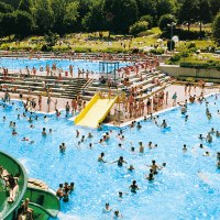 Ein belebtes Freibad mit vielen Menschen im Wasser und auf der Liegewiese. Eine gelbe Rutsche und eine grüne Wasserrutsche sind sichtbar., © Stadt Wendlindgen am Neckar
