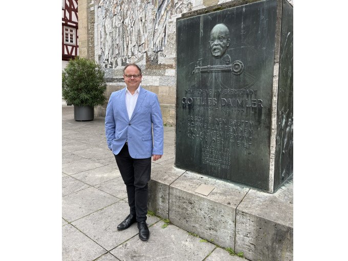 Ein Mann in einem blauen Sakko steht neben einem Denkmal von Gottlieb Daimler in Schorndorf. Im Hintergrund ist ein Wandrelief zu sehen., &copy; Eigenbetrieb Tourismus und Citymanagement Schorndorf