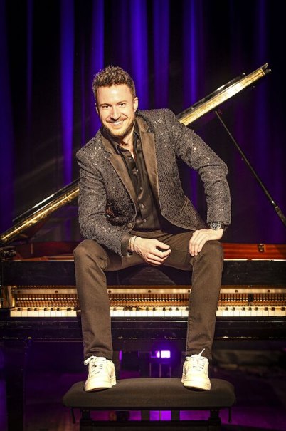 Ein Mann in einem glitzernden Anzug sitzt l&auml;chelnd auf einem Klavier. Der Hintergrund ist in violettem Licht gehalten, was eine B&uuml;hnenatmosph&auml;re schafft., &copy; Stadthalle Leonberg