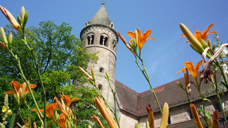 Turm des Klosters Lorch ragt in den blauen Himmel, umgeben von grünen Bäumen und orangefarbenen Lilien im Vordergrund., © Remstal Tourismus e.V.