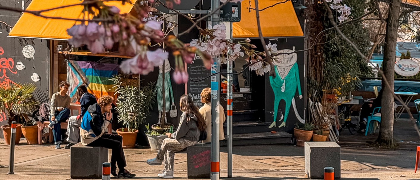 Ein gem&uuml;tliches Stra&szlig;encaf&eacute; mit gelbem Sonnenschutz, Regenbogenflagge und bl&uuml;henden Kirschbl&uuml;ten. Menschen sitzen entspannt davor., &copy; Caf&eacute; Moody, Stuttgart