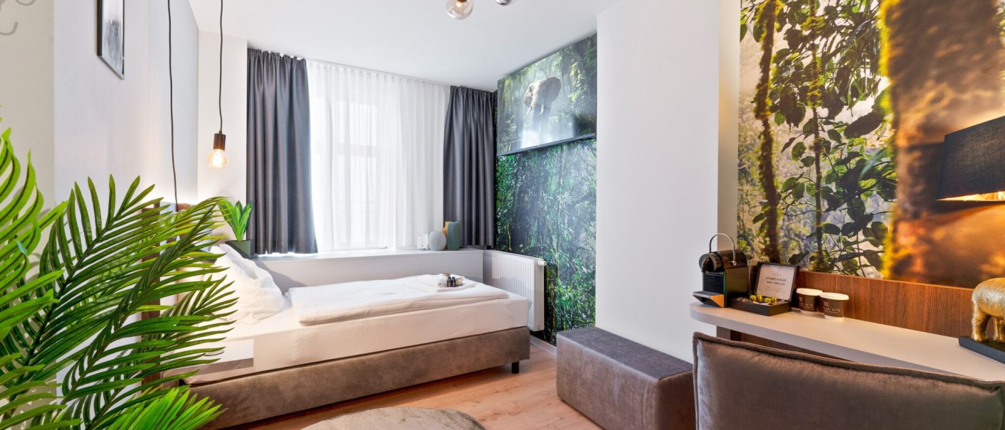 Ein modernes Einzelzimmer mit Dschungelmotiv an der Wand, einem Bett, Schreibtisch und Pflanzen. Helle Beleuchtung und stilvolle Einrichtung., © JUNGLE N°5 GmbH