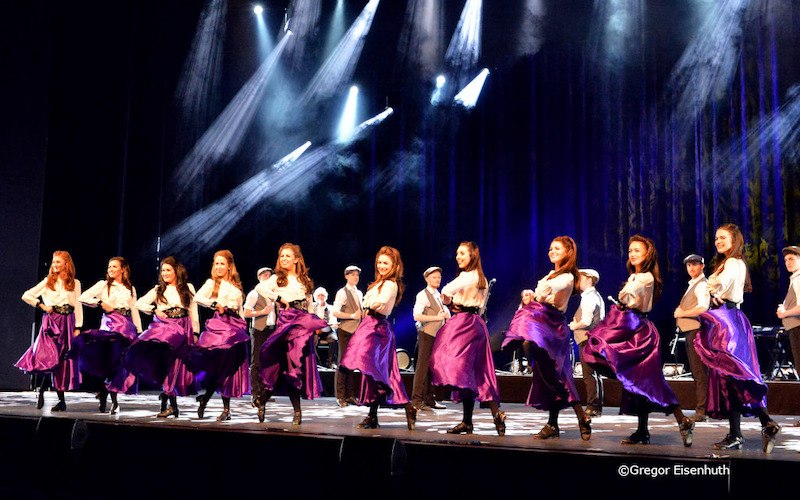 Tanzgruppe in lila Röcken und weißen Blusen auf einer Bühne, beleuchtet von Scheinwerfern. Im Hintergrund sind Musiker zu sehen., © Kultur- und Sportamt Stadt Bietigheim-Bissingen