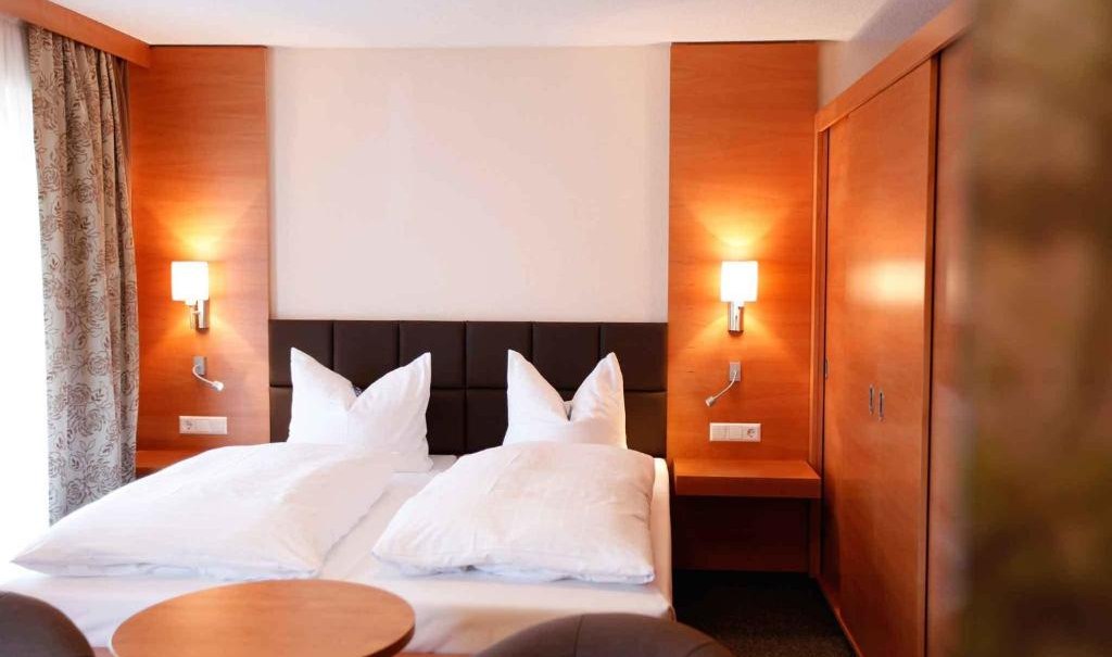 Gemütliches Doppelzimmer mit Holzmöbeln, großem Bett, zwei Nachttischlampen und einem kleinen Tisch mit zwei Sesseln., © Nödinger Gastro GmbH