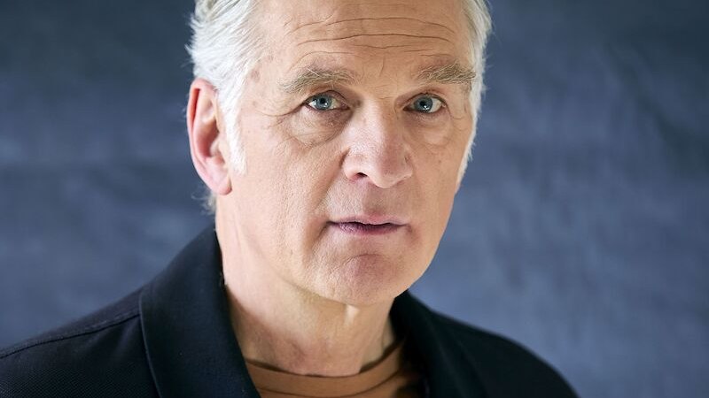 Ein &auml;lterer Mann mit grauem Haar und nachdenklichem Ausdruck blickt in die Kamera. Er tr&auml;gt eine dunkle Jacke und ein braunes Shirt vor einem dunklen Hintergrund., &copy; Stadthalle Leonberg