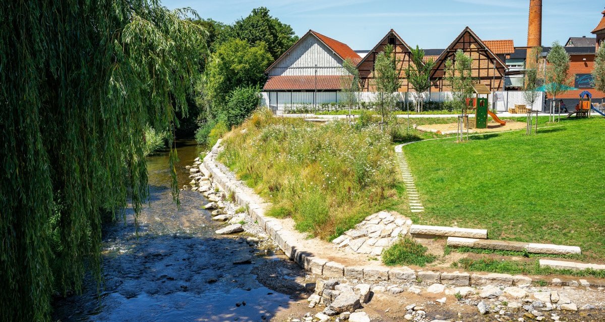 Ein Fluss flie&szlig;t an einem Park mit Spielplatz und Fachwerkh&auml;usern vorbei. Im Hintergrund ist ein hoher Schornstein zu sehen., &copy; Remstal Tourismus e.V.