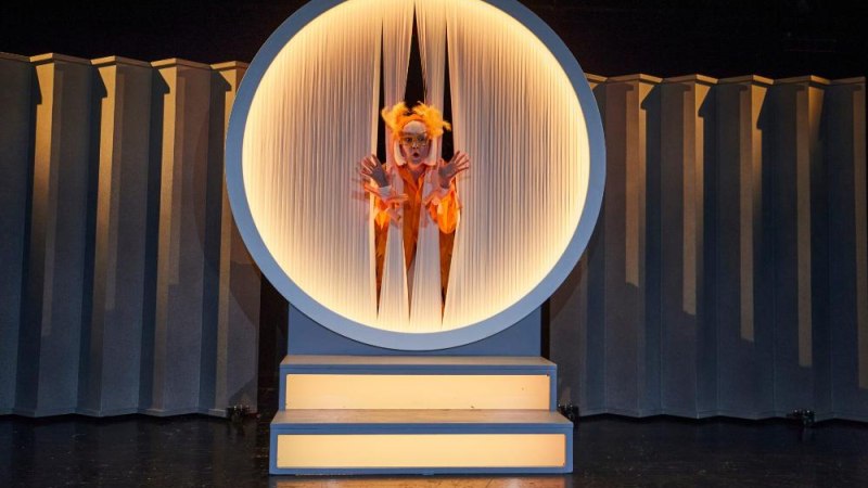 Eine Person in einem orangefarbenen Kostüm schaut durch einen runden, beleuchteten Rahmen auf einer Bühne., © Bernd Eidenmüller