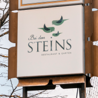 Ein Schild des Restaurants 'Bei den Steins' mit Vögeln als Logo, vor einem kahlen Baum und blauem Himmel., © Stuttgart-Marketing GmbH, Sarah Schmid Ein Schild des Restaurants 'Bei den Steins' mit Vögeln als Logo, vor einem kahlen Baum und blauem Himmel., © Stuttgart-Marketing GmbH, Sarah Schmid