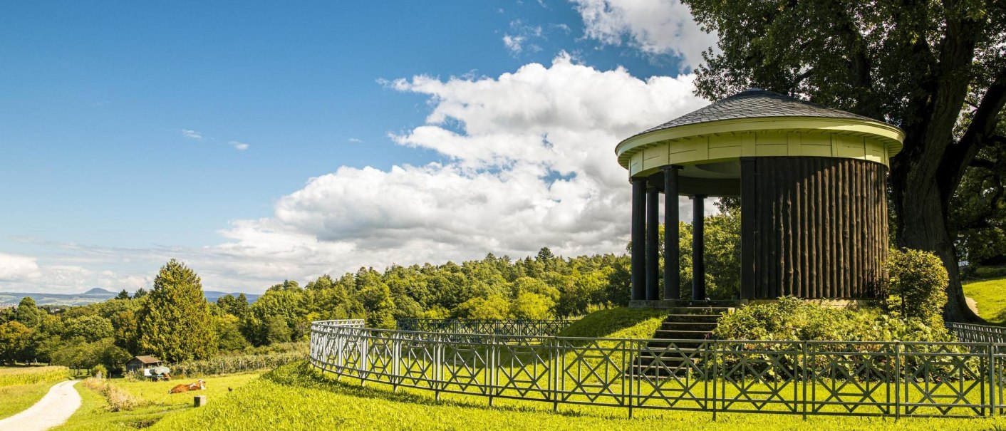 Ein runder Pavillon auf einem H&uuml;gel, umgeben von gr&uuml;ner Landschaft und einem blauen Himmel mit Wolken. Ein Weg f&uuml;hrt durch die Wiese., &copy; SMG, Sarah Schmid