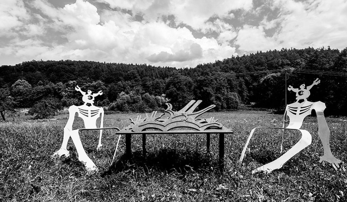 Zwei stilisierte Metallfiguren sitzen auf einer Wiese an einem Tisch mit Buchmotiv. Im Hintergrund erstreckt sich ein Wald unter einem bewölkten Himmel., © Natur.Nah. Schönbuch & Heckengäu Zwei stilisierte Metallfiguren sitzen auf einer Wiese an einem Tisch mit Buchmotiv. Im Hintergrund erstreckt sich ein Wald unter einem bewölkten Himmel., © Natur.Nah. Schönbuch & Heckengäu