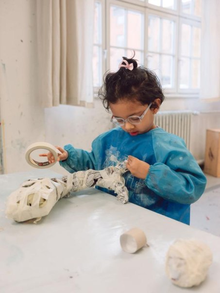 Ein Kind in einem blauen Malkittel bastelt konzentriert mit Klebeband und Papier an einem Tisch in einem hellen Raum., © Kunstschule Labyrinth Ein Kind in einem blauen Malkittel bastelt konzentriert mit Klebeband und Papier an einem Tisch in einem hellen Raum., © Kunstschule Labyrinth