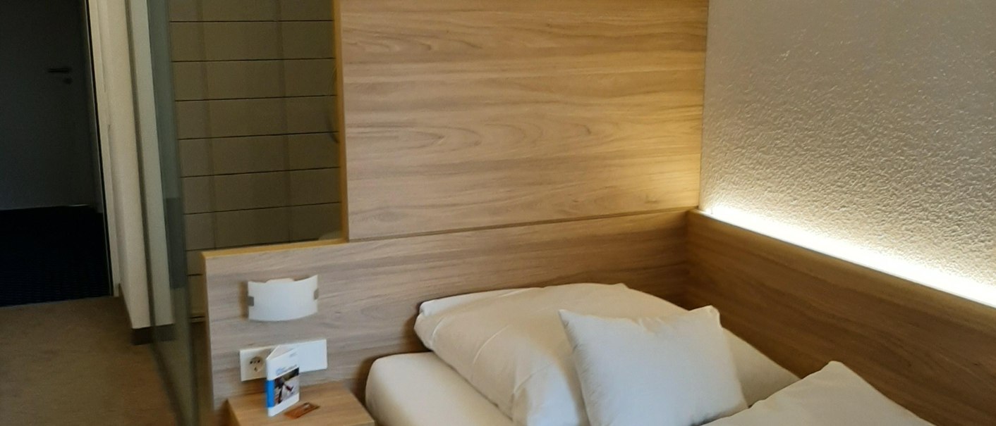 Ein gemütliches Einzelzimmer mit einem Bett, einem kleinen Nachttisch und einer offenen Dusche im Hintergrund., © Hotel am Schlossberg Herrenberg Ein gemütliches Einzelzimmer mit einem Bett, einem kleinen Nachttisch und einer offenen Dusche im Hintergrund., © Hotel am Schlossberg Herrenberg