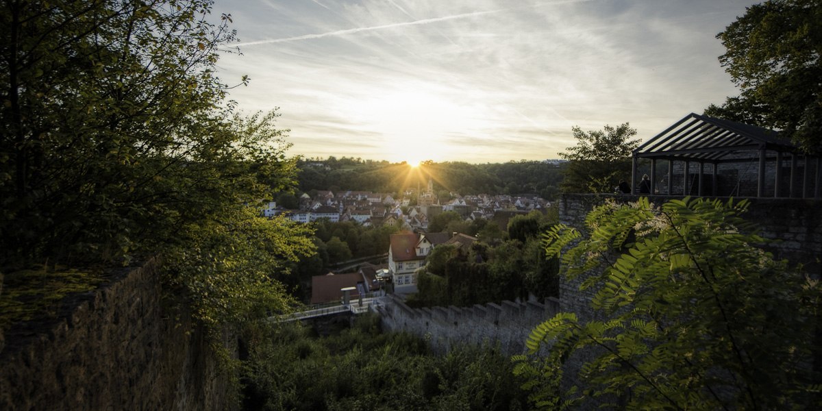 Sonnenuntergang über Schwäbisch Hall, gesehen von einer erhöhten Position mit Bäumen und alten Mauern im Vordergrund., © Patrick Krol