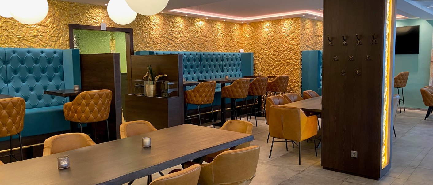 Ein stilvoller Restaurant-Innenraum mit blauen und braunen Sitzm&ouml;beln, goldener Wandverkleidung und runden Deckenleuchten., &copy; Lunar