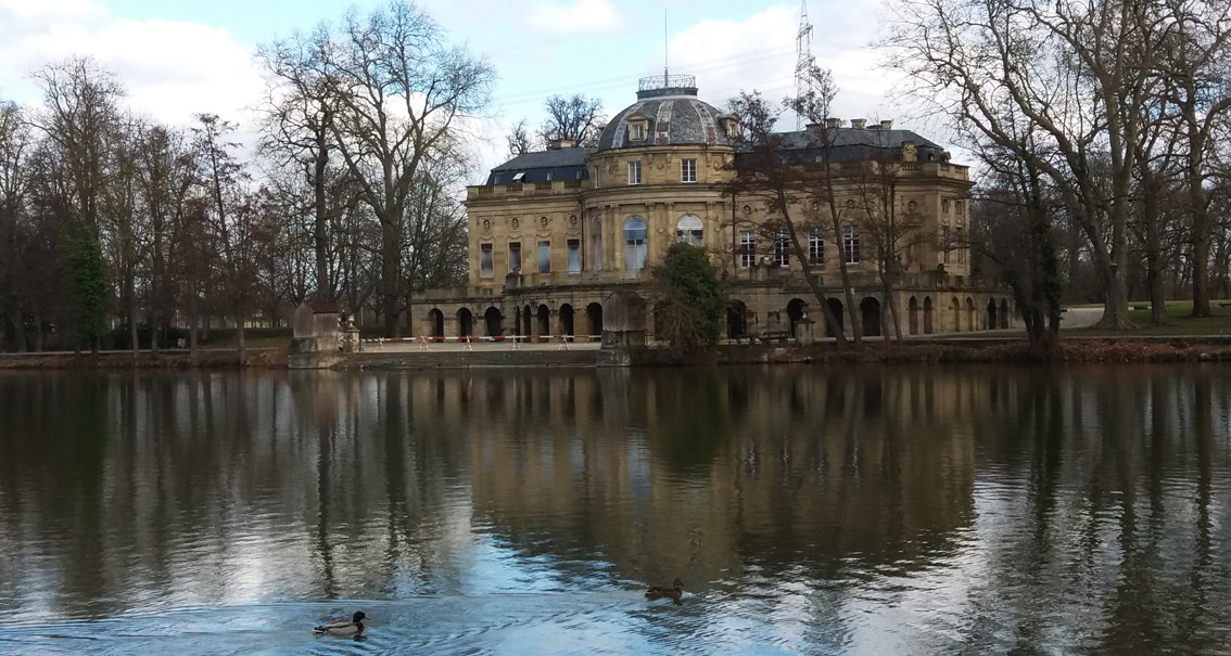 Ein historisches Gebäude spiegelt sich in einem ruhigen See. Im Vordergrund schwimmen Enten. Karge Bäume umgeben das Gebäude., © Flucht-Raum