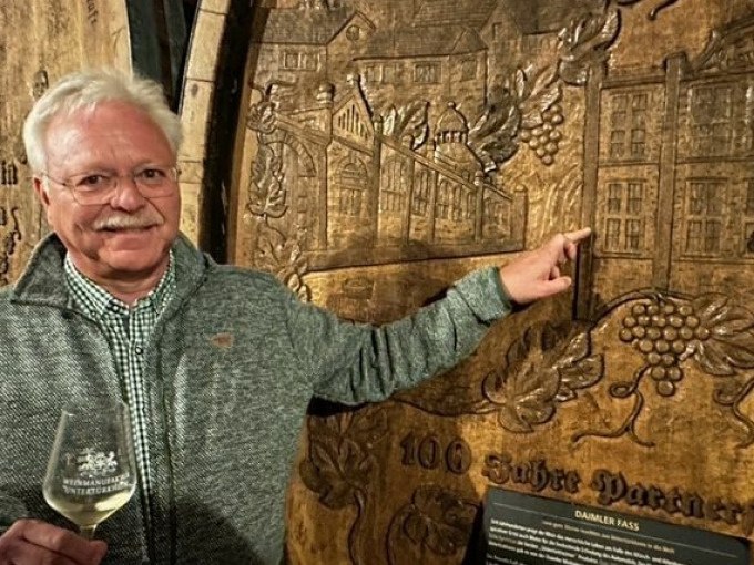 Ein Mann zeigt auf ein kunstvoll geschnitztes Weinfass in einem Weinkeller. Er hält ein Weinglas und lächelt in die Kamera., © Weingenuss Stuttgart Ein Mann zeigt auf ein kunstvoll geschnitztes Weinfass in einem Weinkeller. Er hält ein Weinglas und lächelt in die Kamera., © Weingenuss Stuttgart