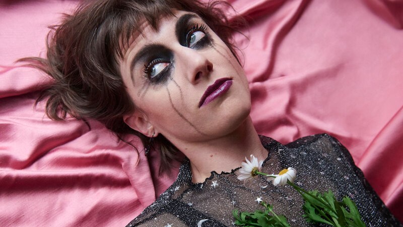 Frau mit starkem Augen-Make-up liegt auf rosa Stoff, trägt ein glitzerndes Oberteil mit Blumen auf der Brust., © Mauerwerk GmbH Frau mit starkem Augen-Make-up liegt auf rosa Stoff, trägt ein glitzerndes Oberteil mit Blumen auf der Brust., © Mauerwerk GmbH