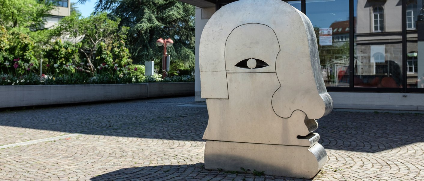 Eine große Metallskulptur in Form eines stilisierten Kopfes steht auf einem gepflasterten Platz, umgeben von Grünpflanzen., © Fotoknobi