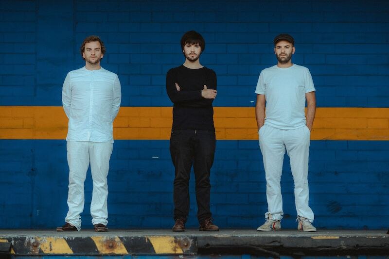 Drei Männer stehen nebeneinander vor einer blau-gelben Wand. Zwei tragen weiße Kleidung, einer schwarze. Sie blicken ernst in die Kamera., © Kulturhaus Schwanen