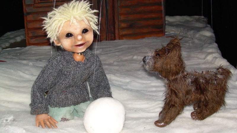 Eine Puppe mit blonden Haaren und grauem Pullover kniet im Schnee neben einem kleinen Hund vor einem Holzhaus., © Zaubermühle Merklingen Eine Puppe mit blonden Haaren und grauem Pullover kniet im Schnee neben einem kleinen Hund vor einem Holzhaus., © Zaubermühle Merklingen