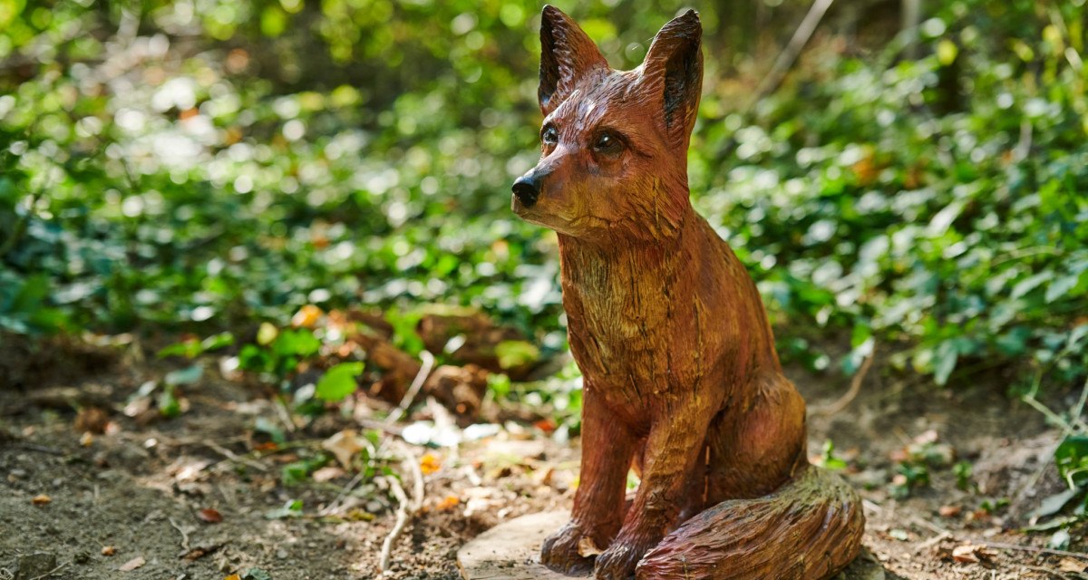 Eine detaillierte Holzskulptur eines Fuchses sitzt auf einem Erlebnispfad im Wald, umgeben von gr&uuml;nem Laub., &copy; Natur.Nah. Sch&ouml;nbuch & Heckeng&auml;u