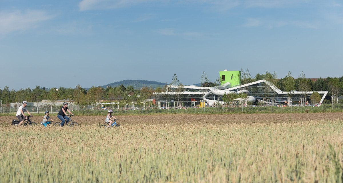 Familie radelt durch ein Feld, modernes Gebäude mit Rutschen im Hintergrund, blauer Himmel., © Stadt Fellbach Familie radelt durch ein Feld, modernes Gebäude mit Rutschen im Hintergrund, blauer Himmel., © Stadt Fellbach
