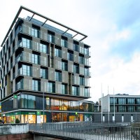 Ein modernes Gebäude mit Glasfassade und Geschäften im Erdgeschoss. Es hat mehrere Stockwerke und eine markante Architektur., © TOMAS