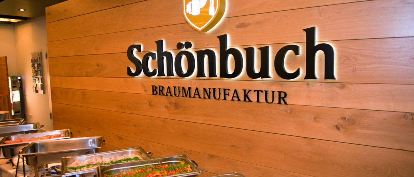 Innenansicht einer Brauerei mit Buffet. An der Holzwand steht "Schönbuch Braumanufaktur". Im Vordergrund sind Speisen in Warmhaltebehältern., © Schönbuchbrauerei, Sebastian Berger