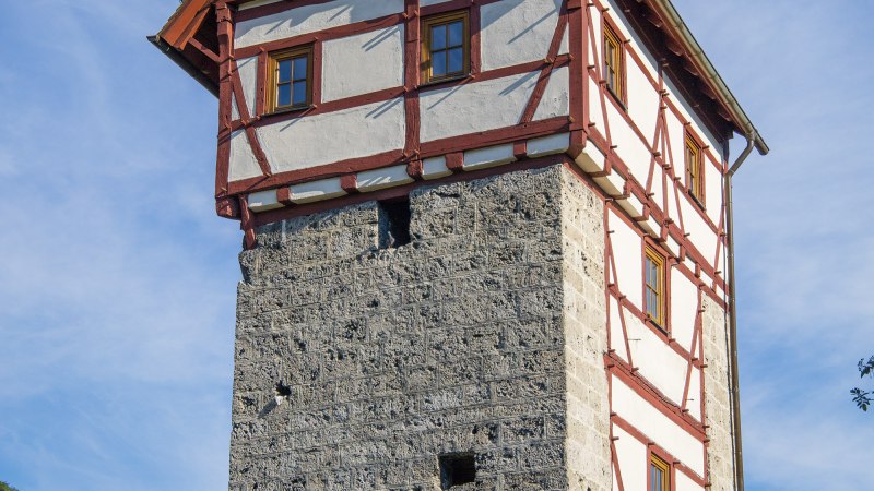 Ein historischer Fachwerkturm in Bad Urach, mit weißem Putz und roten Holzbalken, vor einem klaren blauen Himmel., © Bad Urach Tourismus
